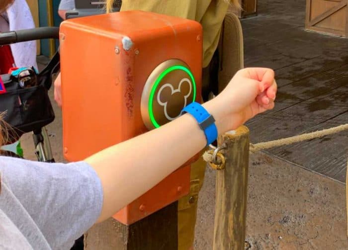 Young girls arm with a blue magic band on it being held next to a box with a green mickey mouse head on it.
