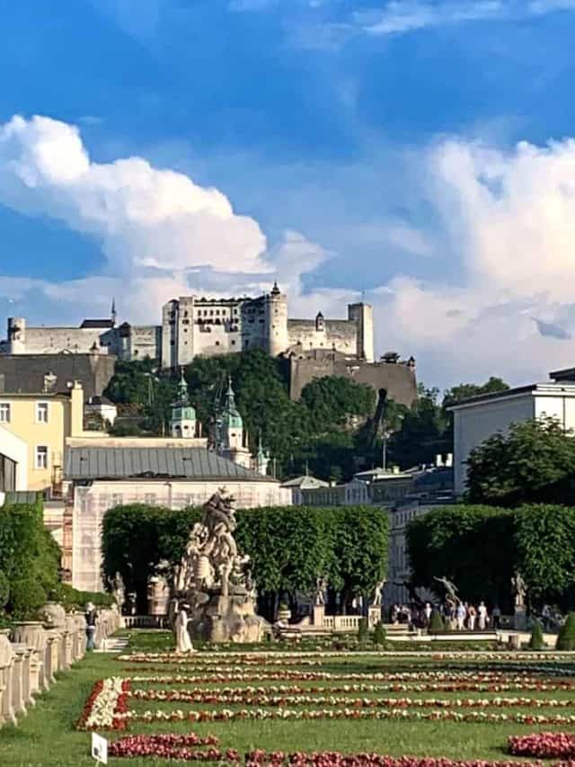 Gardens of red and white flowers amid green grass, statues and water fountains give way to the white Hohensalzburg Fortress at the top of the hill